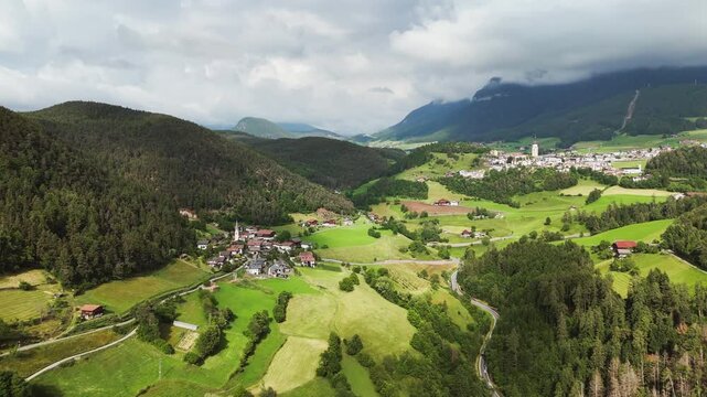 Drone footage of villages across a green mountain valley in South Tyrol, Italy. Alpine countryside beneath low clouds in the Dolomites