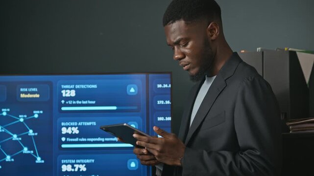 African American young adult software specialist analyzing network data on digital tablet beside cybersecurity monitor. Footage fits corporate software ads and cloud security marketing