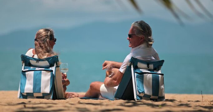 Mature couple enjoy luxury resort vacation, sitting on striped beach chairs with wine glasses, relaxing by the ocean under tropical sun, pointing towards a clear blue sea. Perfect vacation background