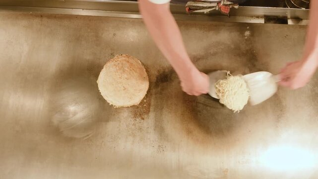Top view of yakisoba noodles dropped and spread on a sizzling iron griddle next to okonomiyaki. Hiroshima style okonomiyaki cooking from above, flat lay, real time 4K.