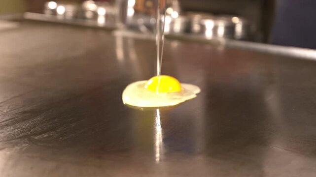 Chef cracks a fresh egg on a hot iron griddle and spreads the yolk with a spatula. Egg layer for Hiroshima style okonomiyaki, sizzling Japanese teppanyaki cooking, real time.