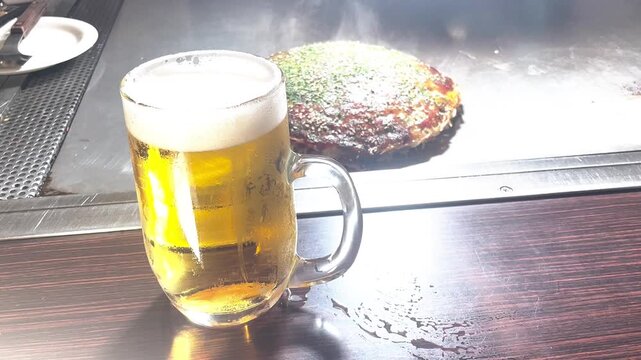 Cold draft beer in a glass mug next to steaming Hiroshima okonomiyaki on a teppanyaki counter. Japanese food and drink, izakaya dinner, appetizing beer and food pairing.