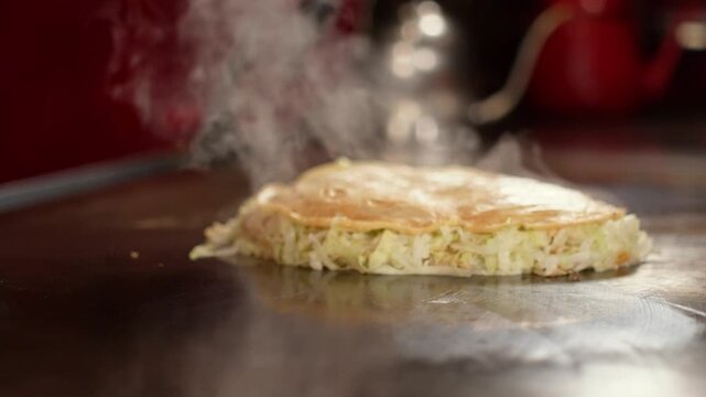 Chef presses Hiroshima okonomiyaki with metal spatulas while steam billows on a hot teppanyaki griddle. Japanese food cooking in a warm red restaurant, static front shot, real time.