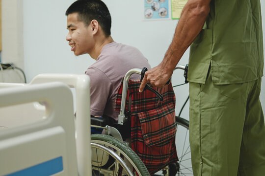 Male nurse in green uniform caregiver pushing disabled young man sitting in wheelchair in hospital ward, healthcare and rehabilitation.