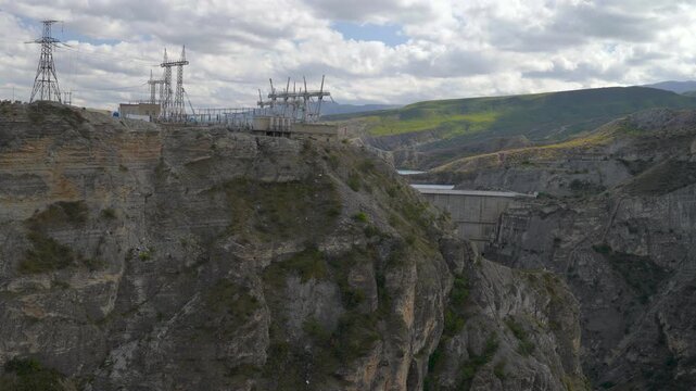 Hydroelectric power facility on steep canyon cliffs with transmission towers, reservoir water, rocky slopes, green hills, and broken cloud cover under wide elevated landscape view