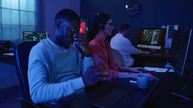 African American young adult man experiencing eye strain while coding in cybersecurity office with multiethnic team. Ideal for burnout campaigns, tech promos and digital health advertising