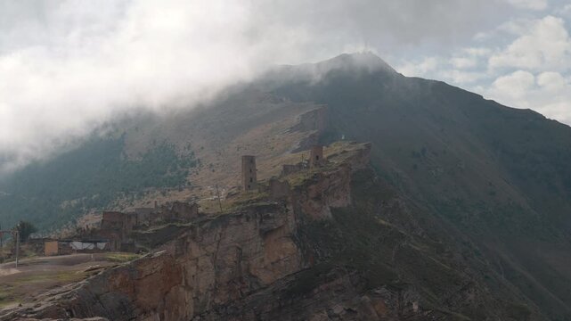 Clouds drift over rugged mountain ridge with weathered cliffside ruins, steep rock layers, sparse vegetation, distant summit, and quiet historic landscape beneath bright hazy sky