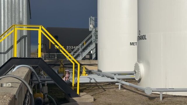 A pan right with a slow push in on a large white methanol storage tank with a yellow railing and pipes leading to it, set against a clear sky.