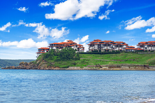 A coastal resort with red-roofed buildings stands on a grassy hill overlooking a calm blue sea under a bright sky with fluffy white clouds.