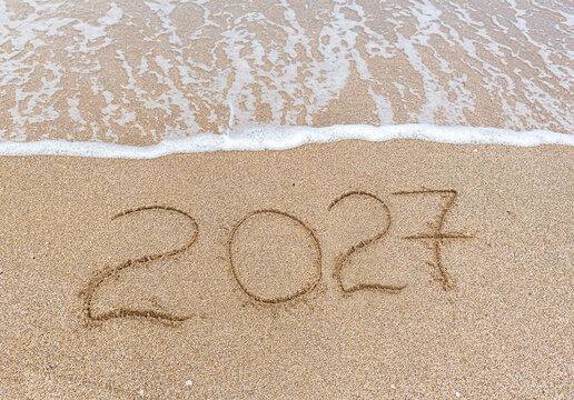 The number 2027 is written in the soft sand near gently washing ocean waves on a sunny beach.