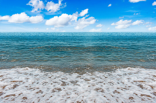 A serene view of blue ocean waves gently washing onto a sandy beach under a bright sky with scattered white clouds.