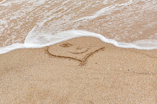 A smiley face icon is drawn in wet sand as gentle ocean waves wash over the sandy beach.