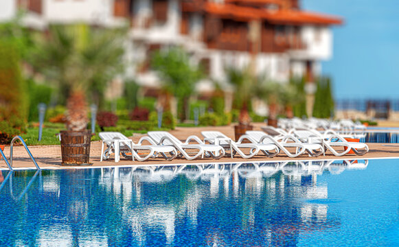 A luxurious swimming pool features clear blue water reflecting white sun loungers under a bright sunny sky.