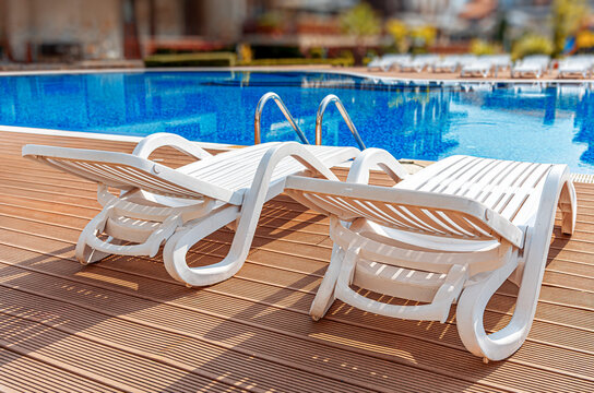 Two white plastic sun loungers sit on a wooden deck next to a clear blue outdoor swimming pool on a sunny day.