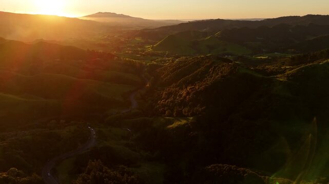 ugged green valley with winding road, dense forest ridges silhouetted against golden sunflare near Raglan, New Zealand, dramatic mountain backdrop at dusk. Aerial drone rising wide shot