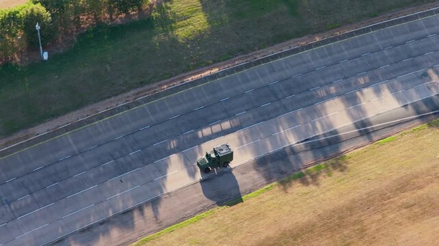 Green tactical vehicle driving along a long straight concrete path at an outdoor automotive testing facility during a sunny afternoon with long shadows cast across the ground