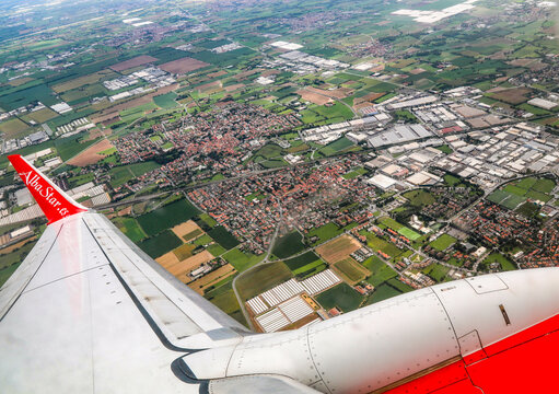 High-altitude aerial view of towns, agricultural fields, and industrial zones seen past the wing and red engine of an AlbaStar airliner.