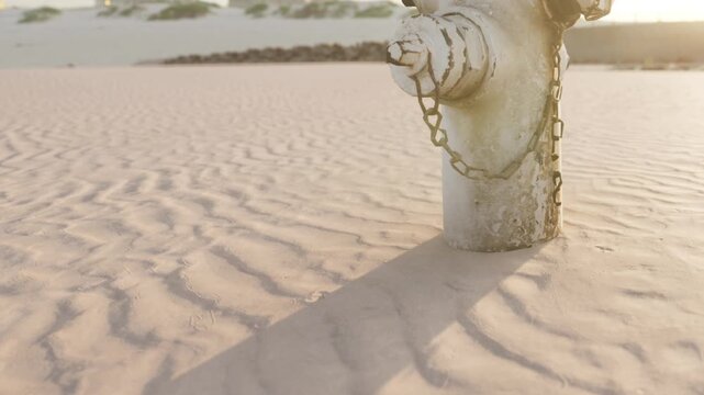 A solitary fire hydrant emerges from the dry sandy ground, casting a long shadow as the sun sets. The surrounding landscape is barren, with subtle ripples in the sand enhancing the serene solitude.