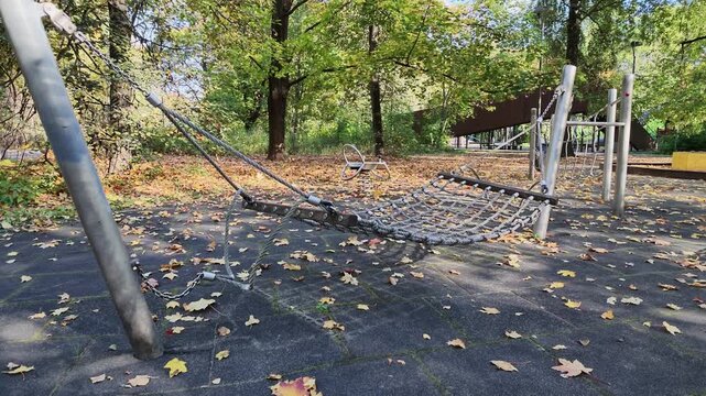 Empty rope net hammock swing between metal posts on an autumn playground with fallen leaves.