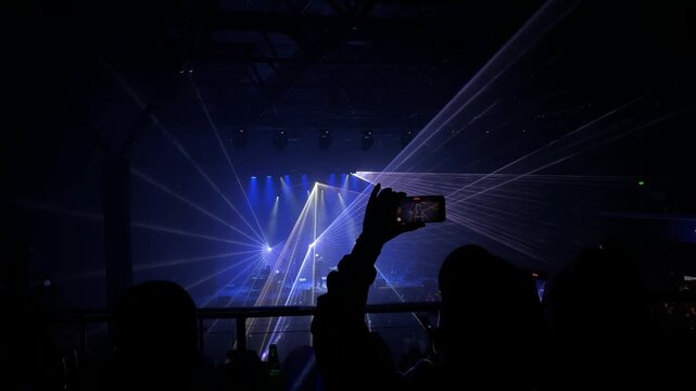 Live music concert in a nightclub with dramatic blue laser lights, on the foreground a man records the performance on a smartphone. The video captures the energetic atmosphere of a modern live music