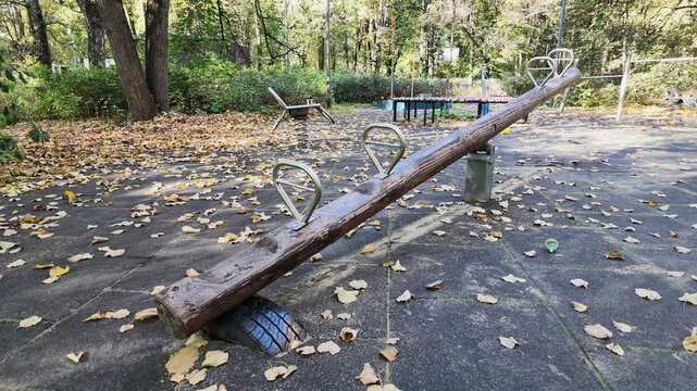 Empty wooden seesaw with metal handles on a playground covered in yellow fallen leaves.