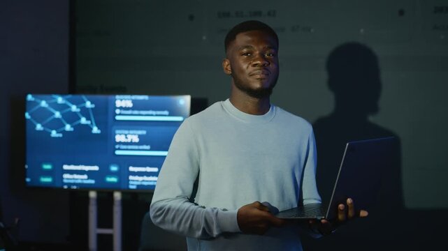 Portrait of young adult African American developer typing on laptop and looking at camera in cybersecurity office. Clip suits tech advertising, cybersecurity, data analytics promo
