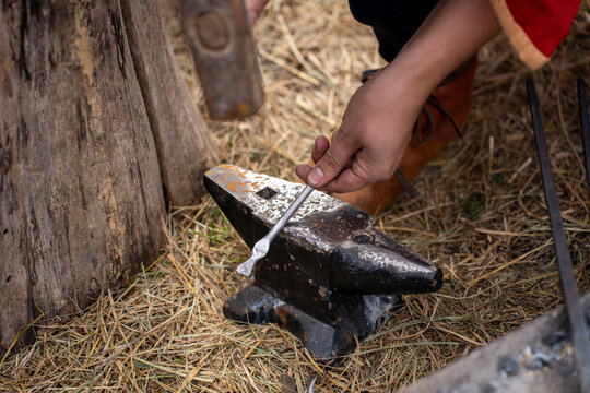 A blacksmith holds a metal rod on a worn anvil in an outdoor work area covered with straw.