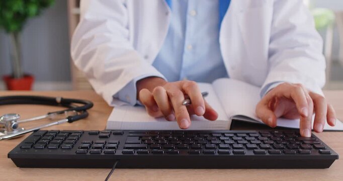 Doctor working at desk in office. Unrecognizable male physician or general practitioner sitting at table, taking notes in notebook, typing on computer keyboard, checking patients' EMRs. Crop closeup
