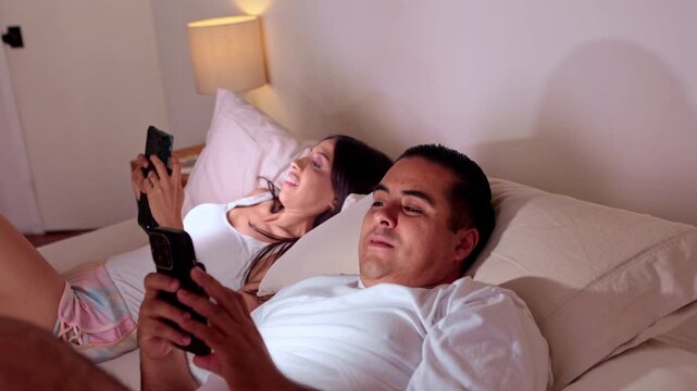 Millennial couple scrolling social media on smartphones in bed at night