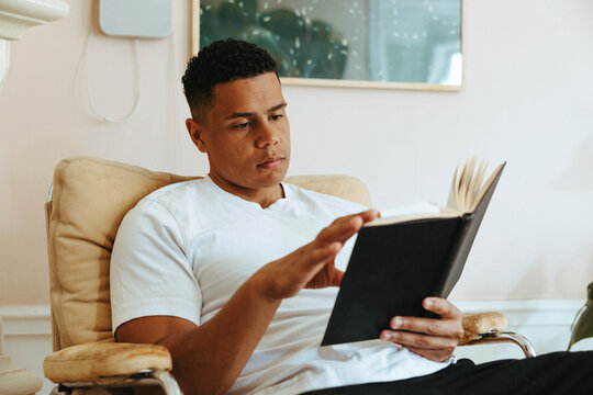 Young man spending leisure time reading book while sitting on armchair at home