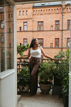 Full length of carefree woman looking away while standing amidst potted plants in balcony at home