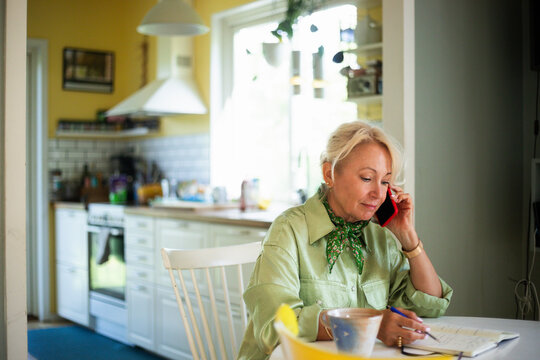 Blond senior woman talking on smart phone while writing in diary at home