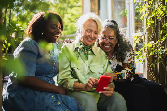 Happy multiracial women sitting beside senior female friend scrolling on smart phone in backyard garden