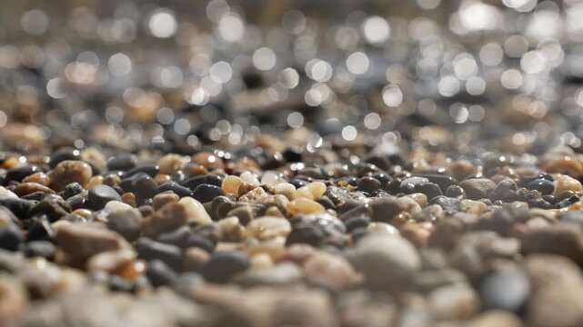 Detailed texture of smooth wet coastal gravel in bright sunlight with shimmering slow motion ocean waves, static macro nature scene