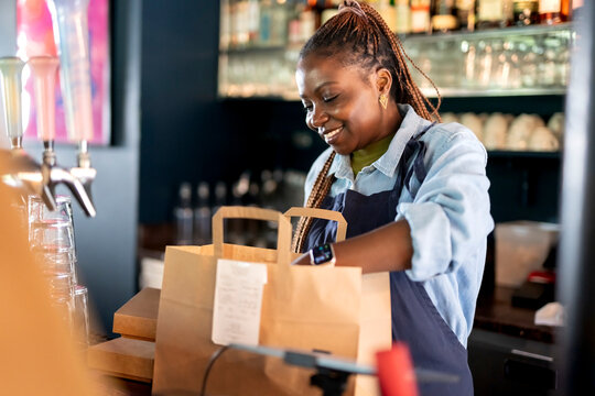 Smiling female owner packing food in paper bags for delivery at restaurant