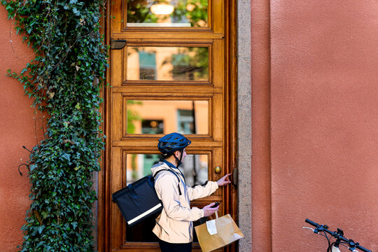 Young delivery man with parcel paper bag ringing intercom of building entrance