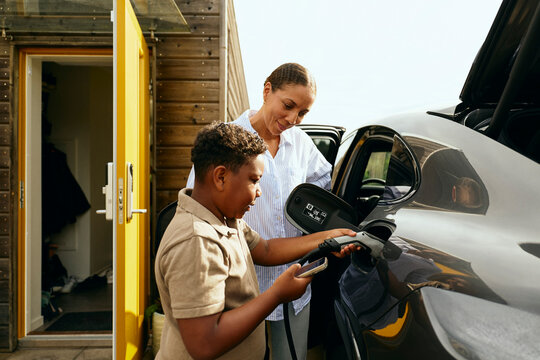 Mother standing beside son plugging connector in electric car port while checking battery on smart phone at backyard