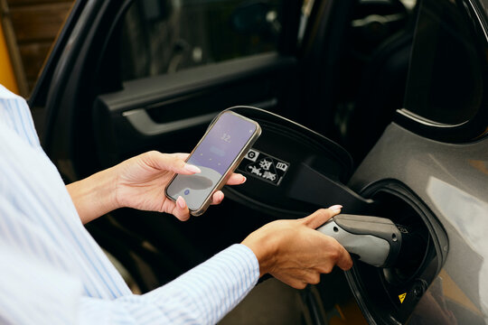 Woman connecting electric plug in charging port of car while checking heating percentage on smart phone
