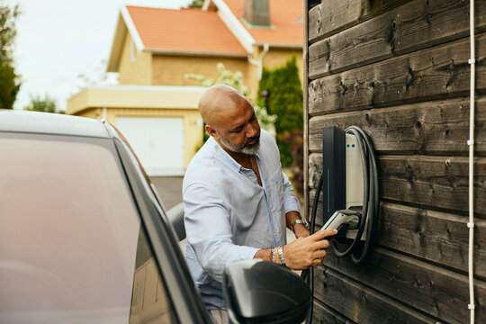 Bald man plugging connector on charging station mounted on outside wall of house