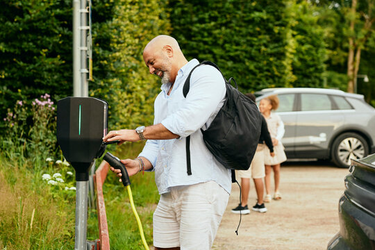 Smiling mature man carrying backpack removing plug from electric vehicle charging pedestal