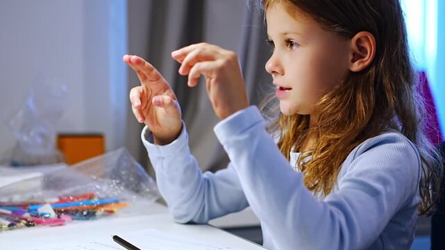 First grade girl stretches tired fingers during handwriting practice at home. Child takes a break from writing to relieve hand fatigue with selective focus