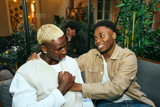 Smiling young man consoling his worried best friend during social gathering in greenhouse