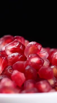 Peeled pomegranate fruit arils in a white bowl slowly rotating against a black background.