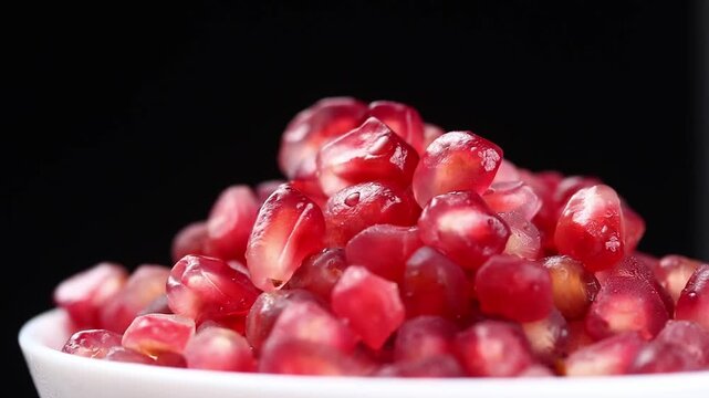 Peeled pomegranate fruit arils in a white bowl slowly rotating against a black background.