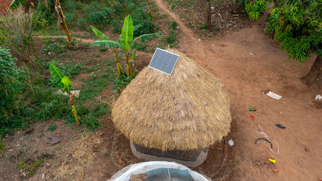 Aerial view of a traditional thatched-roof hut with a modern solar panel installed on top, surrounded by dirt paths and banana trees in Mbale, Eastern Region, Uganda.