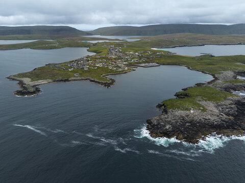Aerial view of Fugla Ness Lighthouse and the coastal village of Hamnavoe surrounded by rocky cliffs, green hills, and the sea in Hamnavoe, Scotland, United Kingdom.