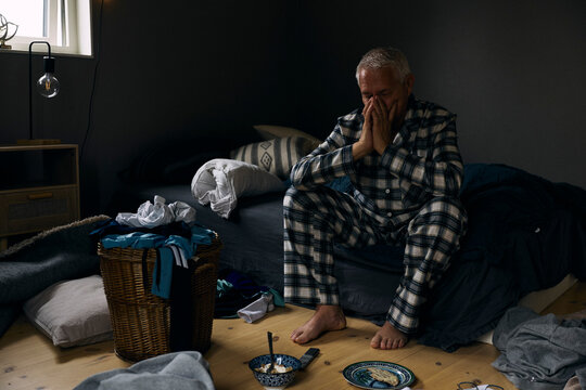 Full length of stressed senior man sitting on bed in messy bedroom at home