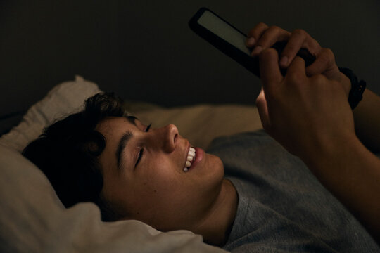 Smiling teenage boy spending leisure time using smart phone in bed at night