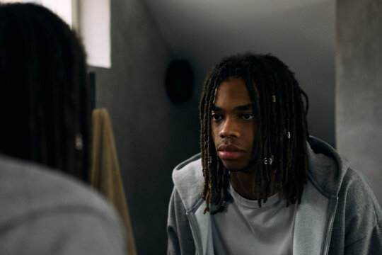 Young man with dreadlocks looking at his reflection in bathroom mirror