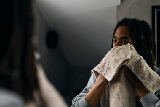 Young man wiping his face with towel while looking at his reflection in mirror